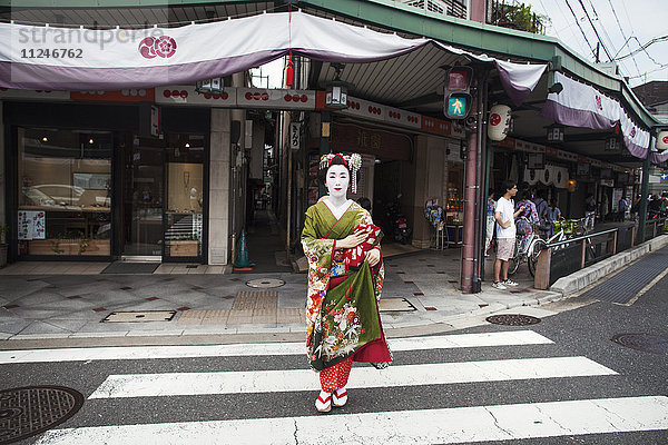 Eine im traditionellen Geisha-Stil gekleidete Frau  in Kimono und Obi  mit aufwändiger Frisur und blumigen Haarspangen  mit weißer Gesichtsschminke mit leuchtend roten Lippen und dunklen Augen  die eine Straße überquert.