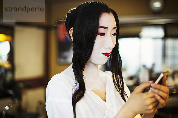 Geisha mit langen schwarzen Haaren und traditionellem weißen Gesichts-Make-up mit einem Smartphone.
