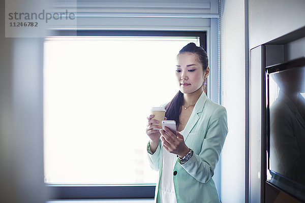 Eine Geschäftsfrau  die sich auf die Arbeit vorbereitet  eine Tasse Kaffee hält und ihr Smartphone kontrolliert.