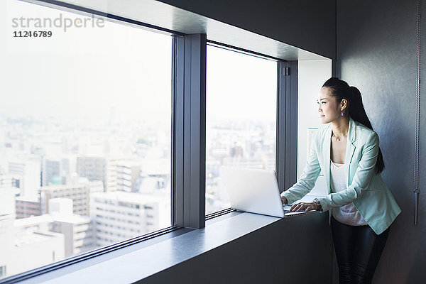 Eine Geschäftsfrau am Fenster mit Blick über die Stadt  die nach draußen blickt. Ein Laptop.