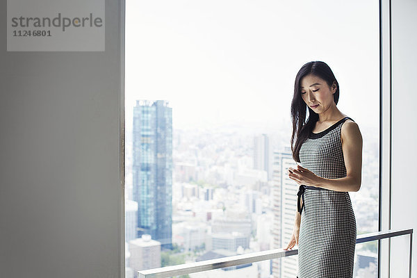 Eine Geschäftsfrau am Fenster mit Blick über die Stadt  die ihr Smartphone benutzt.