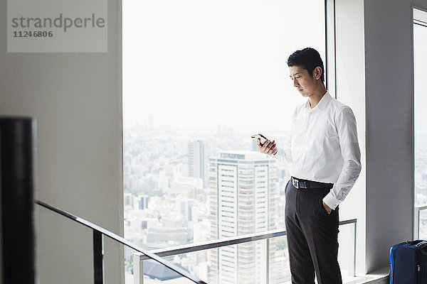 Ein Geschäftsmann im Büro  an einem großen Fenster  schaut auf sein Smartphone.