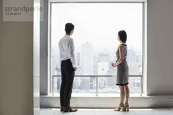 Ein Geschäftsmann und eine Geschäftsfrau stehen an einem großen Fenster mit Blick auf eine Stadt.