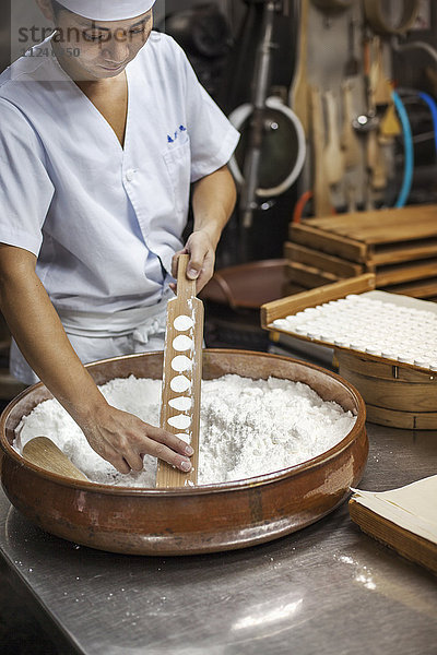Ein kleiner Handwerker  der Wagashi herstellt. Ein Mann  der in einer Großküche eine große Schüssel mit Zutaten mischt und den gemischten Teig in Formen presst.