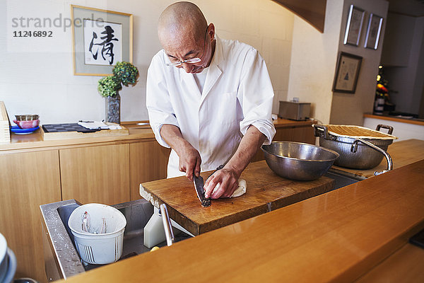 Ein Koch  der in einer kleinen Großküche arbeitet  ein Itamae oder Meisterkoch  der Fisch mit einem großen Messer in Scheiben schneidet  um Sushi zuzubereiten
