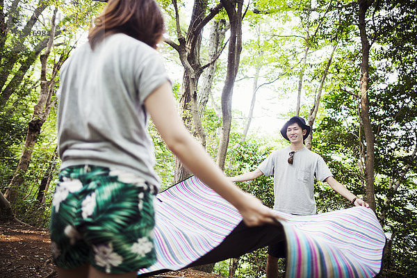 Junge Frau und Mann stehen in einem Wald und halten einen Picknickteppich in der Hand.