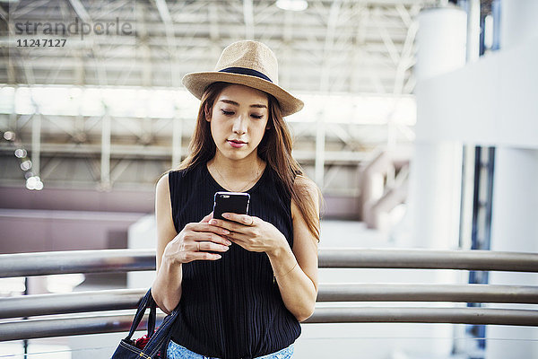 Junge Frau mit langen braunen Haaren  trägt einen Panamahut  in einem Einkaufszentrum  mit einem Mobiltelefon.