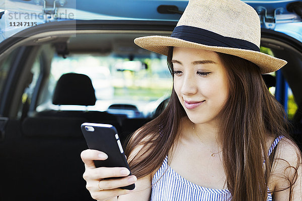 Lächelnde junge Frau mit langen braunen Haaren  die einen Panamahut trägt und ein Mobiltelefon benutzt.