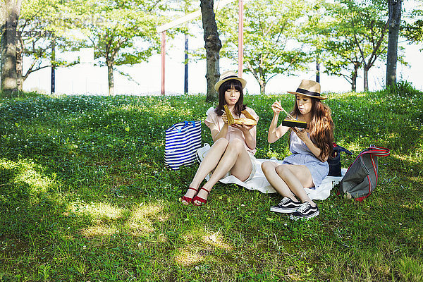 Zwei junge Frauen mit langen braunen Haaren sitzen auf einem Rasen.
