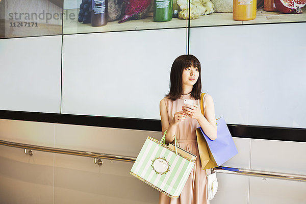 Junge Frau mit langen braunen Haaren  die ein Mobiltelefon und Einkaufstaschen hält.