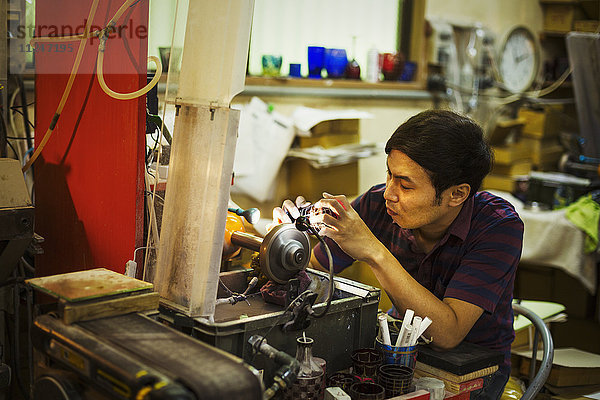 Ein Handwerker sitzt bei der Arbeit und benutzt eine Schleifmaschine  um ein Muster auf einen Glasgegenstand zu ätzen und zu markieren.