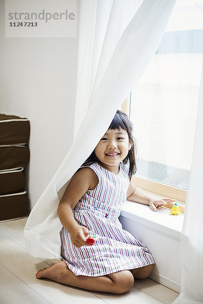 Familienhaus. Ein Mädchen spielt an einem Fenster und versteckt sich hinter dem Vorhang.