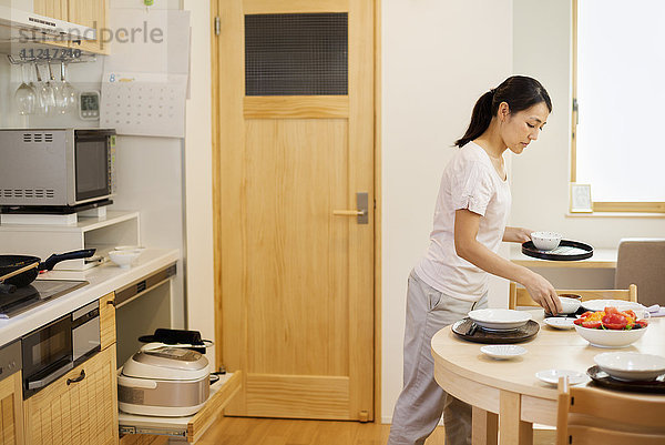 Familienhaus. Eine Frau  die in einer Küche eine Mahlzeit zubereitet und Geschirr auf den Tisch stellt.