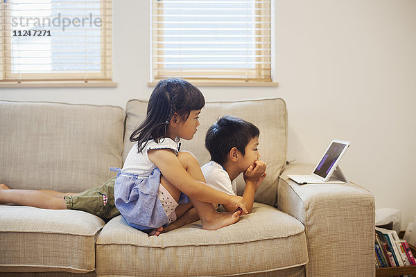 Familienhaus. Ein Junge und ein Mädchen liegen auf einem Sofa und schauen sich ein digitales Tablet an.