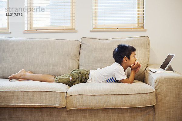 Familienhaus. Ein Junge liegt auf einem Sofa und betrachtet ein digitales Tablet.