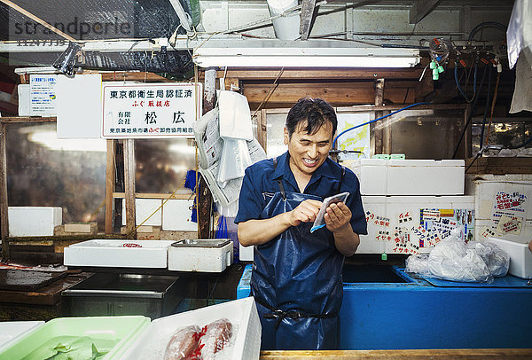 Ein traditioneller Frischfischmarkt in Tokio. Ein Mann in einer blauen Schürze steht hinter der Theke seines Standes und benutzt ein Smartphone  das mit schützendem Plastik überzogen ist.