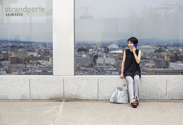 Eine Frau  die an einem Fenster mit Blick auf eine große Stadt sitzt  mit dem Rücken zur Aussicht  telefoniert mit einem Smartphone.