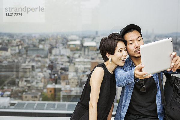 Zwei Menschen  ein Mann und eine Frau  die sich mit einem digitalen Tablett vor dem Blick über eine große Stadt ein Selfie machen.