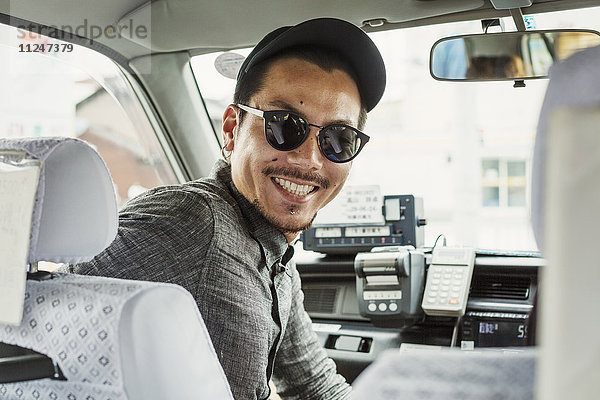 Ein Mann mit Sonnenbrille und Baseballmütze auf dem Beifahrersitz eines Autos dreht sich um und lächelt.