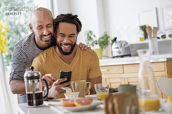 Ein lächelndes homosexuelles Paar benutzt ein Smartphone am Esstisch