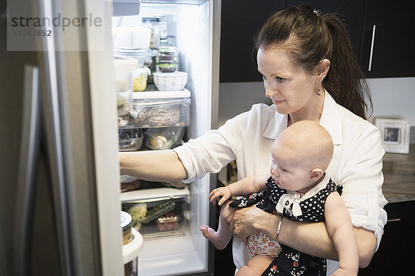Mutter sucht im Kühlschrank nach Essen  während sie ihre kleine Tochter hält (2-5 Monate)