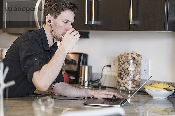Mann trinkt  während er in der Küche am Laptop arbeitet