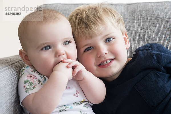 Junge (2-3) mit kleiner Schwester (12-17 Monate) auf dem Sofa sitzend
