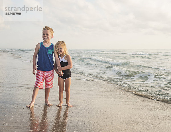 Junge (6-7) und Mädchen (4-5) stehen am Strand am Wasser und halten sich an den Händen