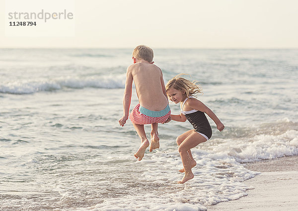 Junge (6-7) und Mädchen (4-5) springen am Strand ins Wasser