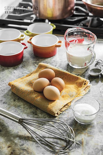 Eier  Milch und Zucker mit Schneebesen und Backförmchen neben Gasherd