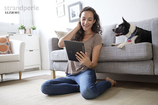 Junge Frau im Wohnzimmer  die ein digitales Tablet mit ihrem Hund benutzt