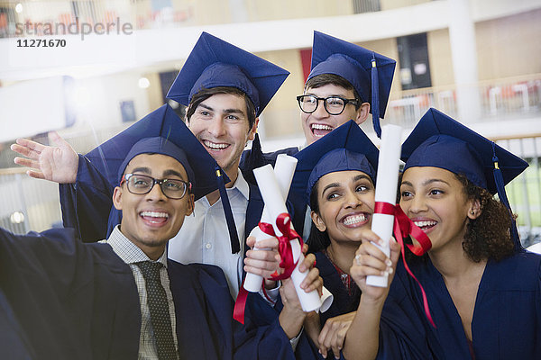 Glückliche Studenten in Mütze und Kleid mit Diplomen  die für Selfie posieren.