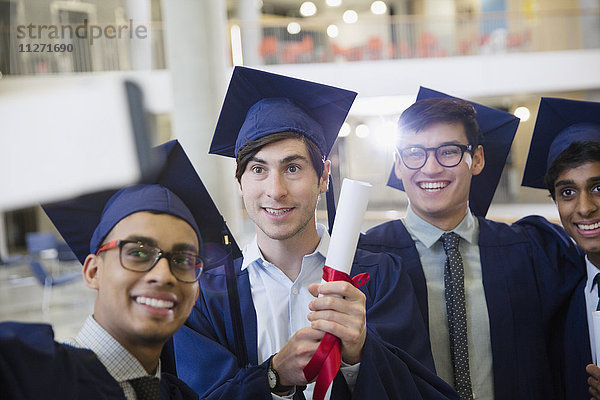 Männliche Hochschulabsolventen in Mütze und Kittel mit Diplom als Selfie