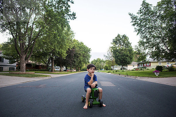 Nachdenklicher Junge gemischter Rasse sitzt auf einem Spielzeugtraktor auf der Straße