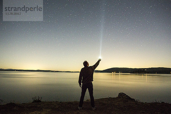 Kaukasischer Mann zeigt mit einer Taschenlampe auf den Nachthimmel in der Nähe von Wasser