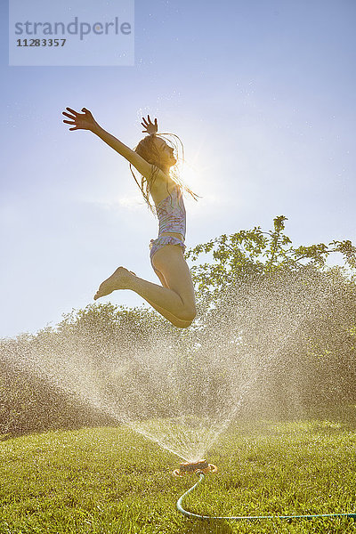 Kaukasisches Mädchen springt über Sprinkleranlage im Garten