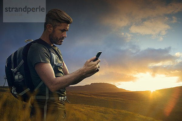 Kaukasischer Wanderer  der bei Sonnenuntergang eine SMS auf seinem Mobiltelefon schreibt