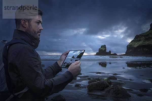 Kaukasischer Wanderer  der bei Sonnenuntergang den Strand mit einem digitalen Tablet fotografiert