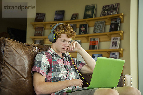 Kaukasischer Junge sitzt auf dem Sofa und hört mit Kopfhörern auf seinen Laptop