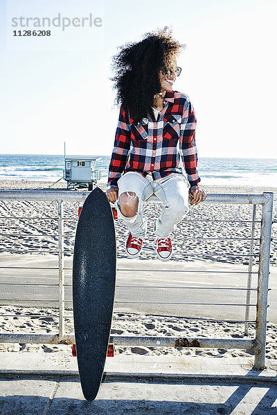 Hispanische Frau sitzt mit Skateboard auf dem Zaun am Strand