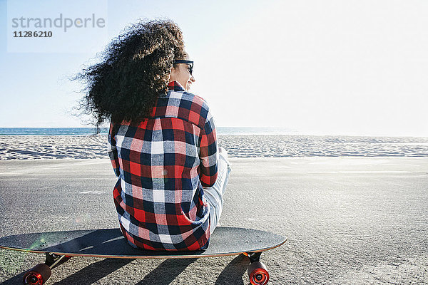 Hispanische Frau sitzt auf einem Skateboard am Strand