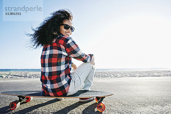 Hispanische Frau sitzt auf einem Skateboard am Strand