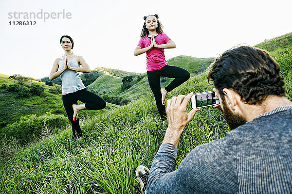 Mann fotografiert Frau und Tochter  die auf einem Hügel Yoga üben