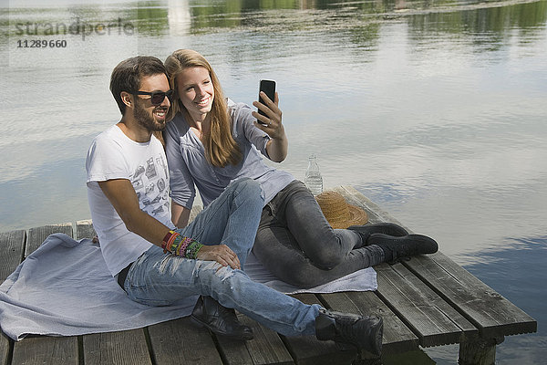 Lächelndes junges Paar sitzt auf einem Steg am See und nimmt einen Selfie.
