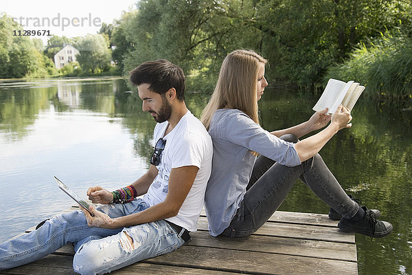 Junges Paar am Steg am See mit Buch und Tablette