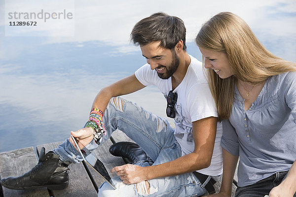 Lächelndes junges Paar sitzt auf einem Steg am See mit Tablette
