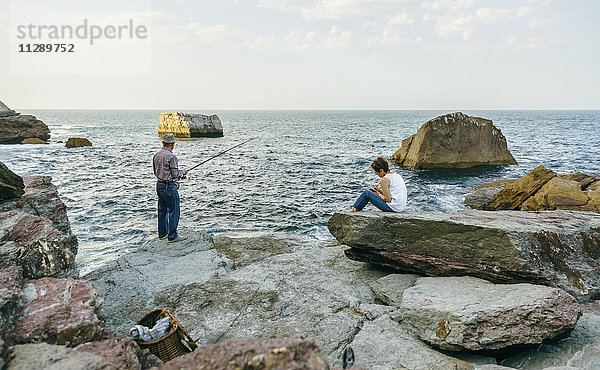 Älterer Mann beim Fischen am Meer mit Frau auf Felsen sitzend
