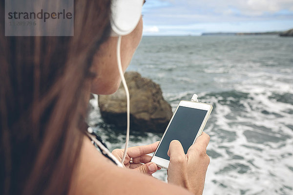 Frau mit Smartphone beim Hören mit Kopfhörern vor dem Meer