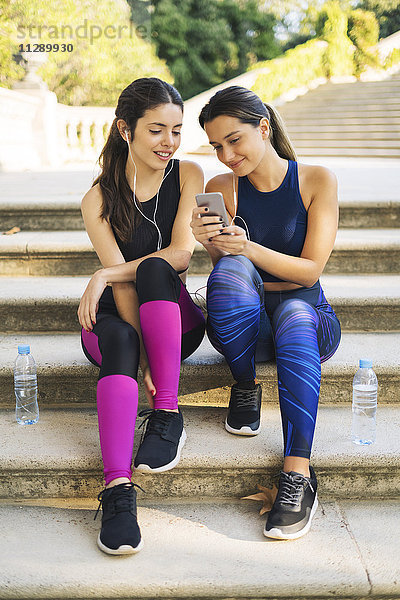 Zwei sportliche junge Frauen sitzen auf der Treppe und schauen aufs Handy.