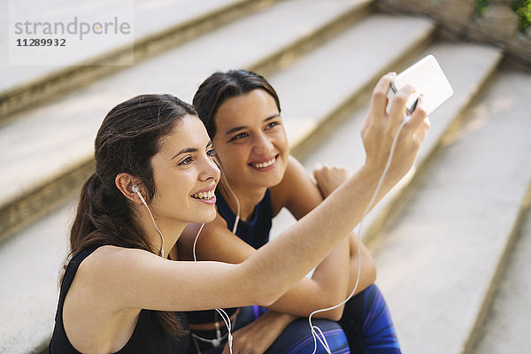 Zwei sportliche junge Frauen sitzen auf einer Treppe und nehmen einen Selfie.